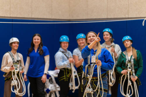 A group of students look up at the recreational therapy ropes course, wearing helmets and holding ropes.